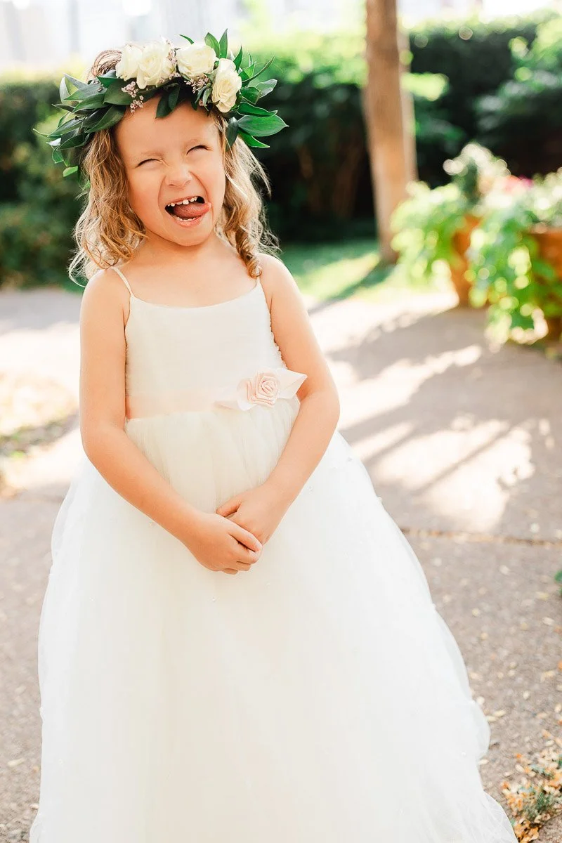 Young girl in a white dress with a floral headpiece smiles widely, standing on a sunlit garden path. The setting exudes joy and innocence.