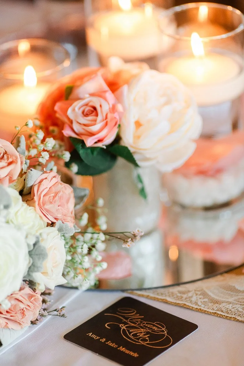 Soft candlelight illuminates a bouquet of pink and white roses with baby's breath on a table. A card with elegant script rests nearby, creating a romantic ambiance.