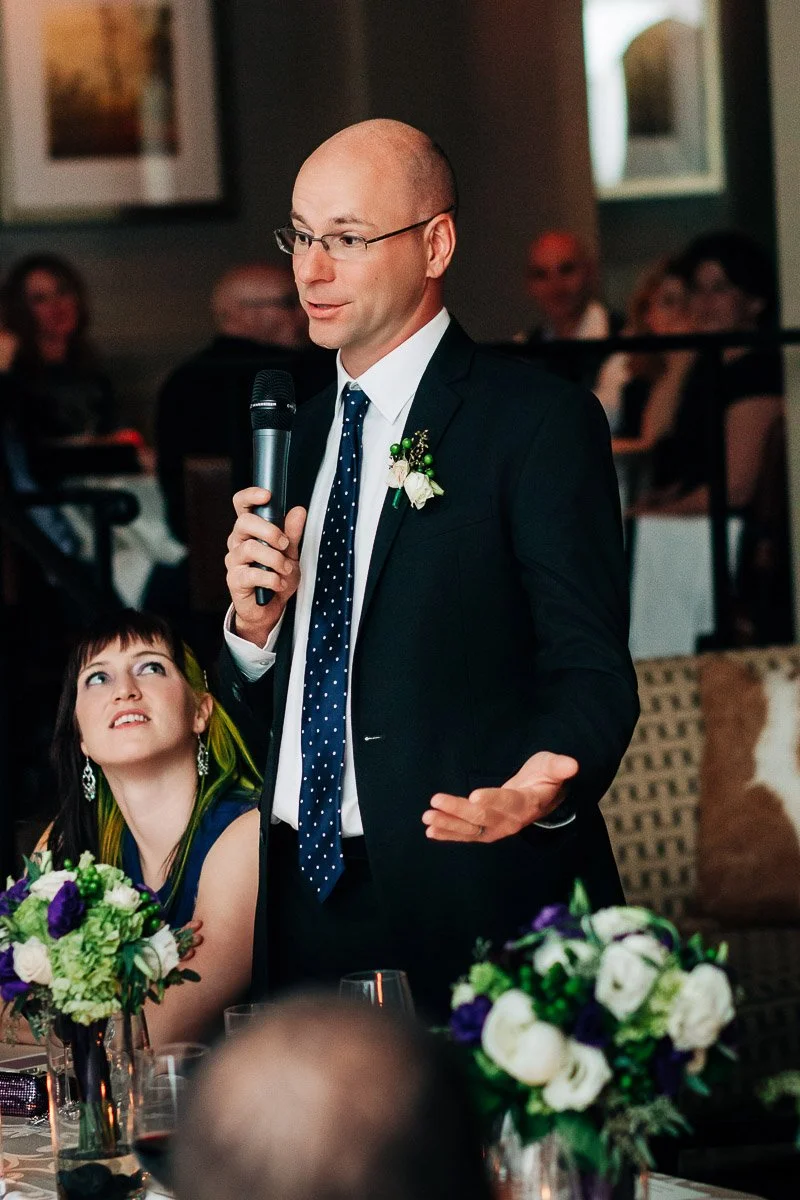 A man in a suit, speaking into a microphone with a confident expression at a formal event. A woman seated with an attentive look. Flowers adorn the table.