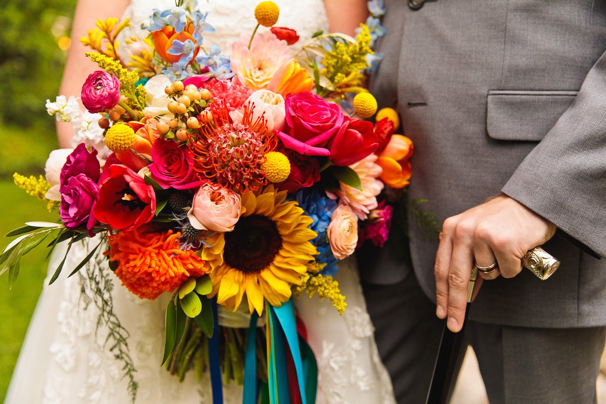 A vibrant bouquet with sunflowers, roses, and mixed blooms held by a bride in lace next to a groom in a gray suit holding a cane. Colorful and joyful.