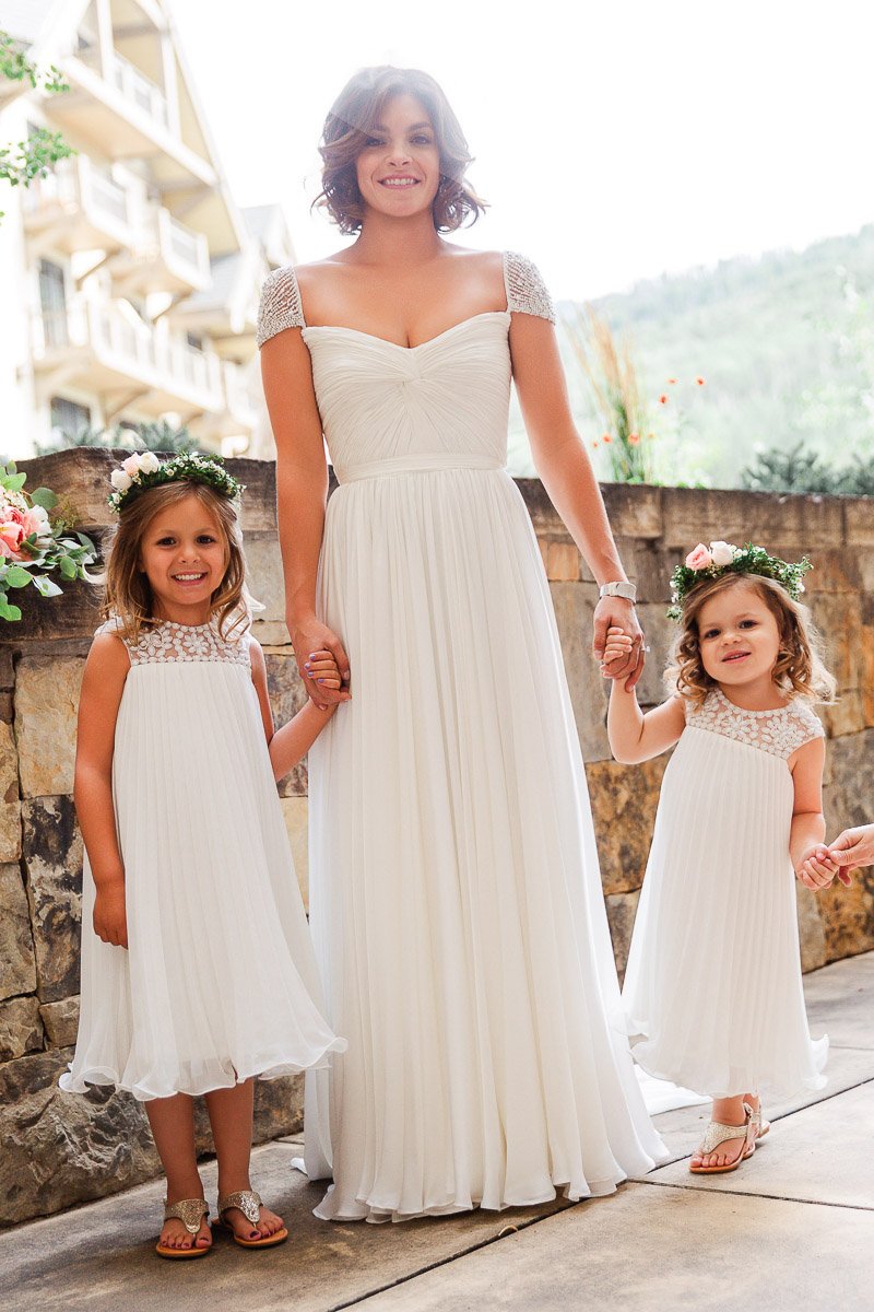 A woman in a white gown stands between two young girls in matching white dresses. They smile, holding hands, with floral crowns, exuding joy.