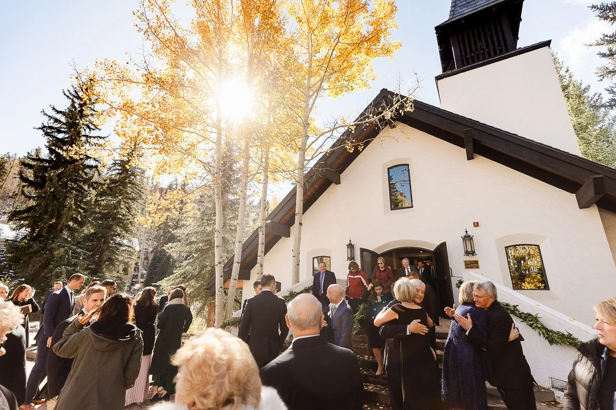 A sunlit scene outside a white chapel with a dark roof, surrounded by autumn trees. Guests in formal attire are joyfully greeting and hugging each other.