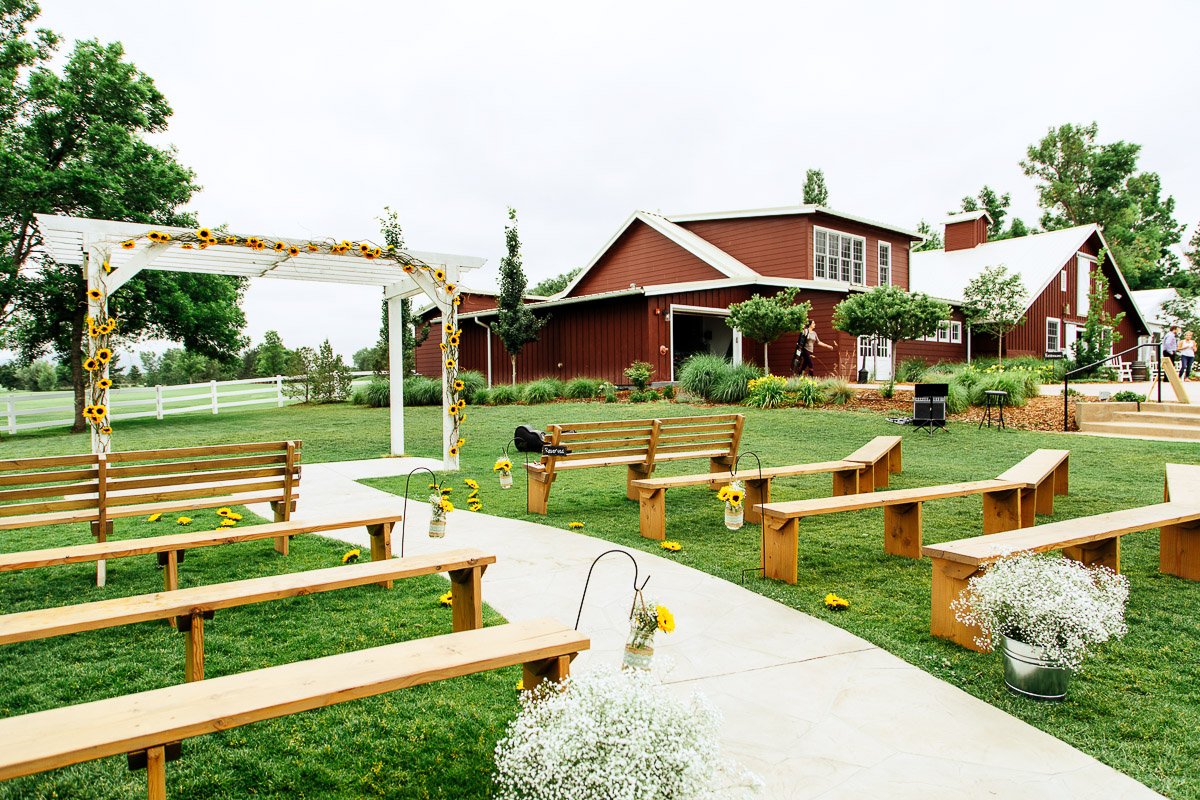 Outdoor wedding setup with wooden benches and a sunflower-decorated archway, set on lush grass in front of a rustic red barn on a cloudy day.