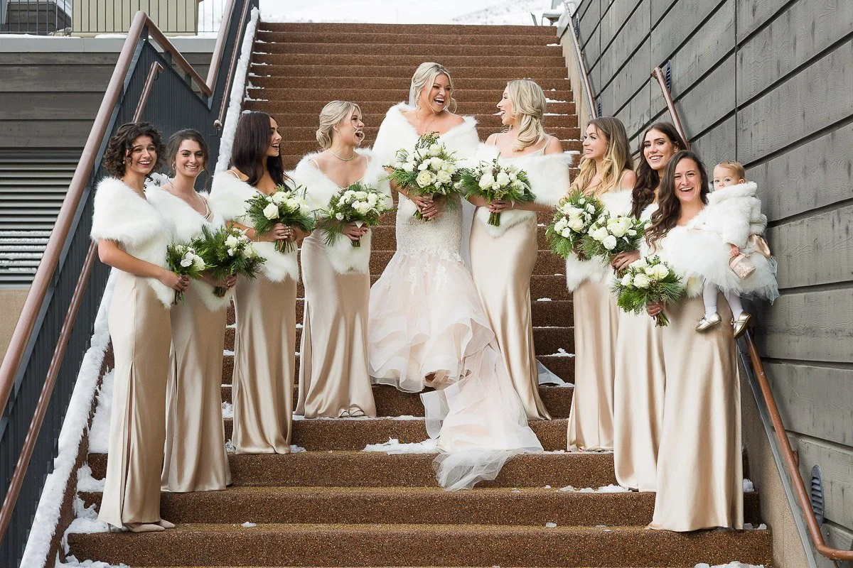 A bride and eight bridesmaids in elegant beige dresses with white fur wraps stand on snowy steps. They hold bouquets, smiling and joyful.