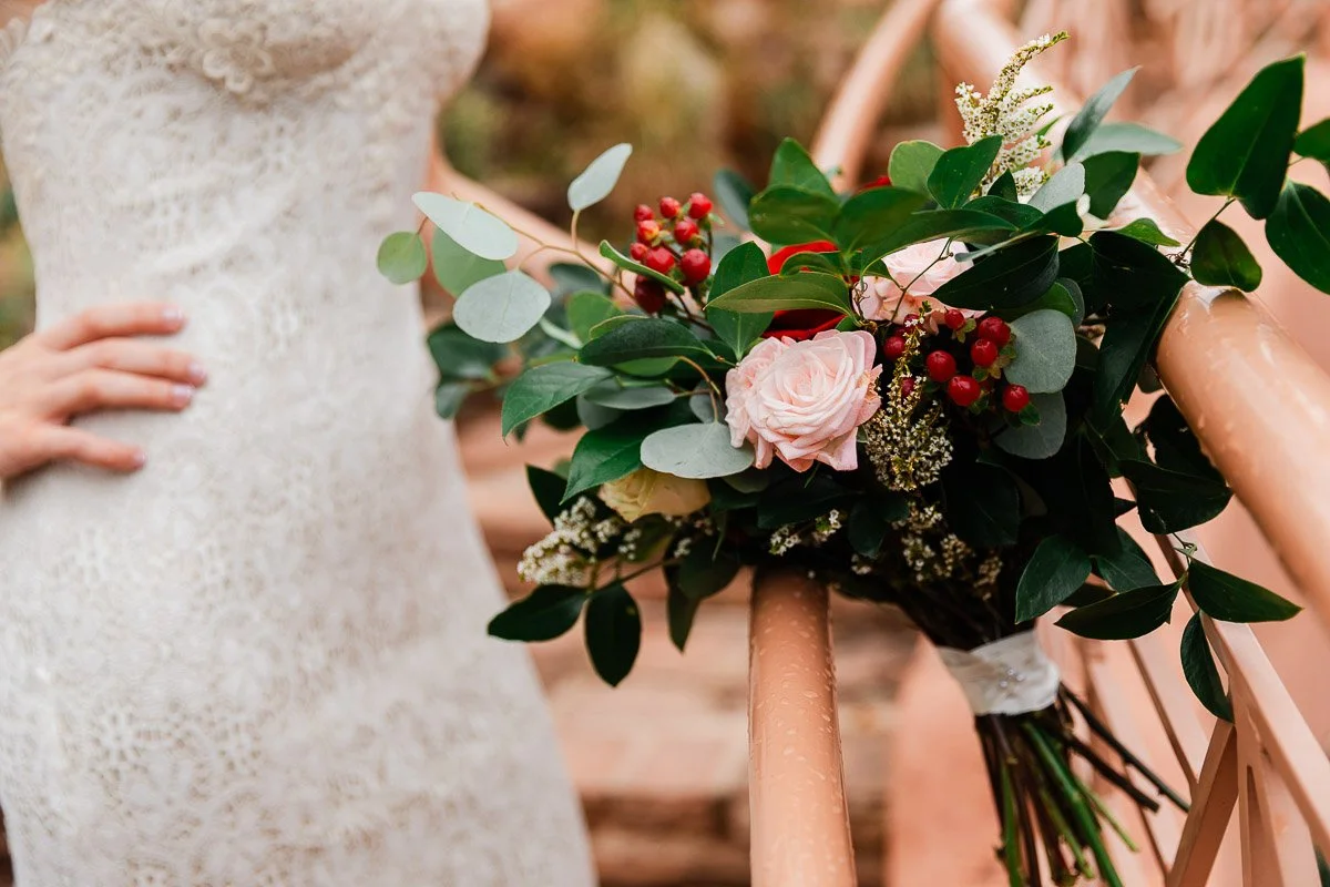 A close-up of a bride's lace dress and a vibrant bouquet resting on a railing, featuring pink roses, red berries, and lush greenery, evoking elegance.