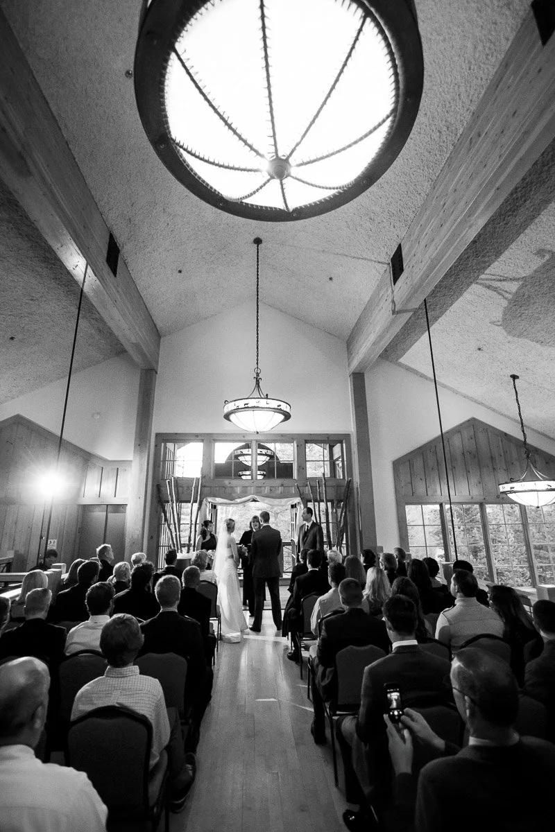 Black and white photo of a wedding ceremony in a tall, wooden-beamed hall. The couple stands facing the officiant, surrounded by seated guests. Elegant and serene.