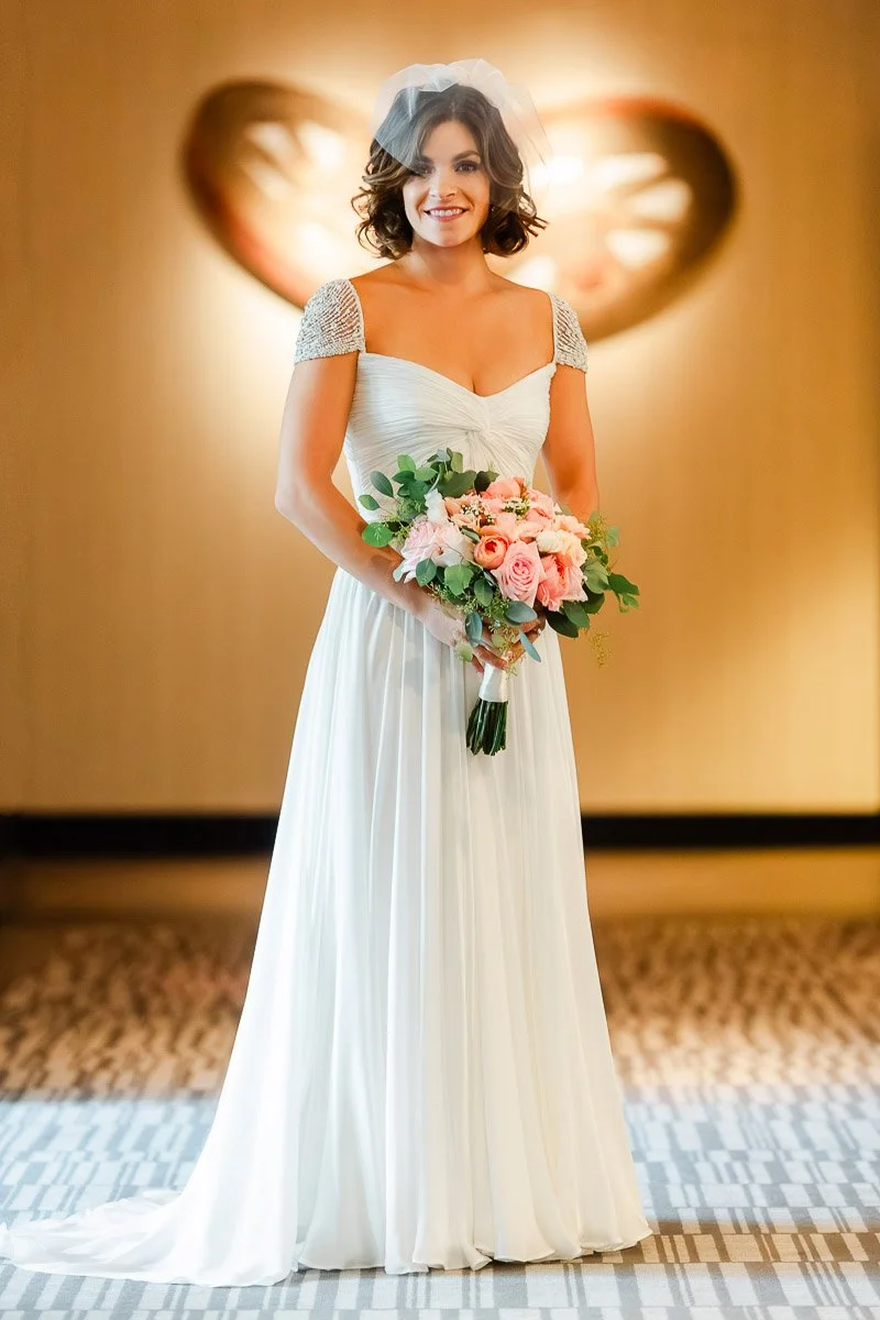 A bride in a flowing white gown and veil smiles, holding a bouquet of pink and white flowers. A heart-shaped decoration is blurred in the background during a Four Seasons Resort wedding in Vail, Colorado