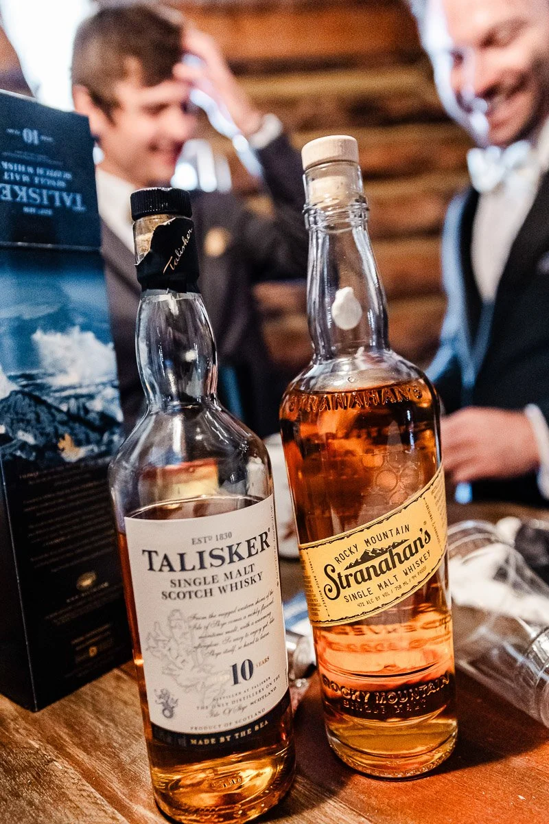 Two bottles of whiskey, Talisker and Stranahan's, are on a wooden table. Two smiling people in suits are in the blurred background, creating a warm, celebratory atmosphere.