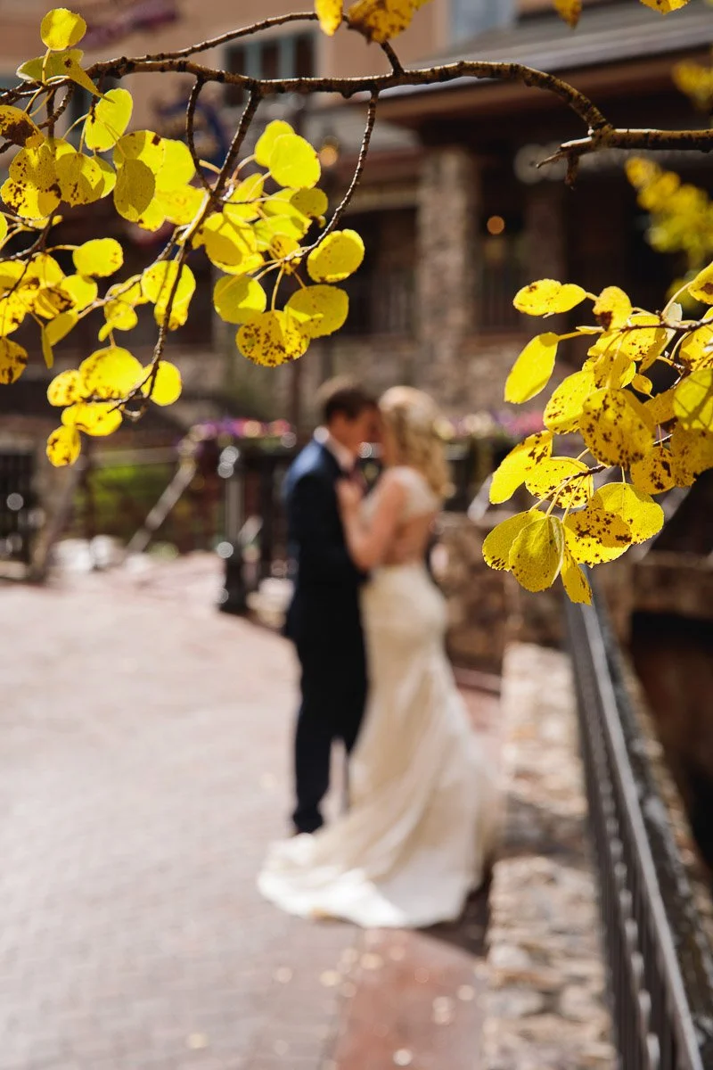 A couple embraces in the background of a softly focused image, framed by vibrant yellow leaves in the foreground, conveying romance and autumn warmth.