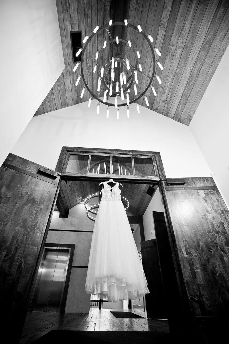 Black and white photo of a wedding dress hanging in a rustic room with a wooden vaulted ceiling and a modern chandelier, creating an elegant ambiance.