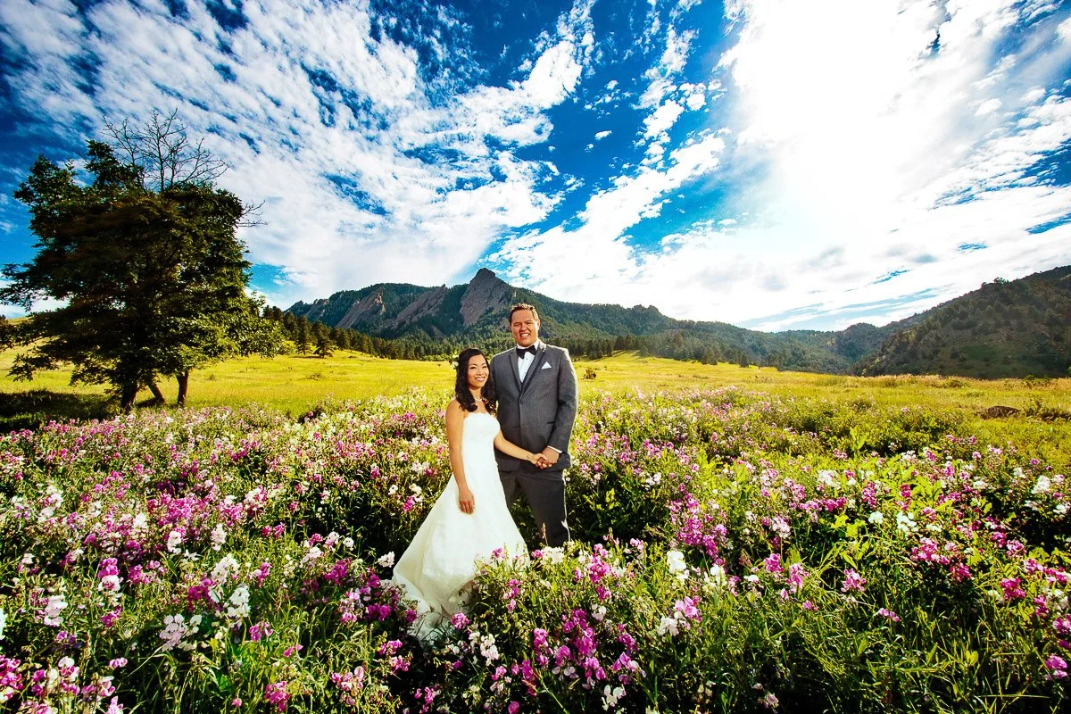 A bride in a white dress and groom in a gray suit stand smiling in a vibrant field of purple and white flowers, with mountains and a bright blue sky.