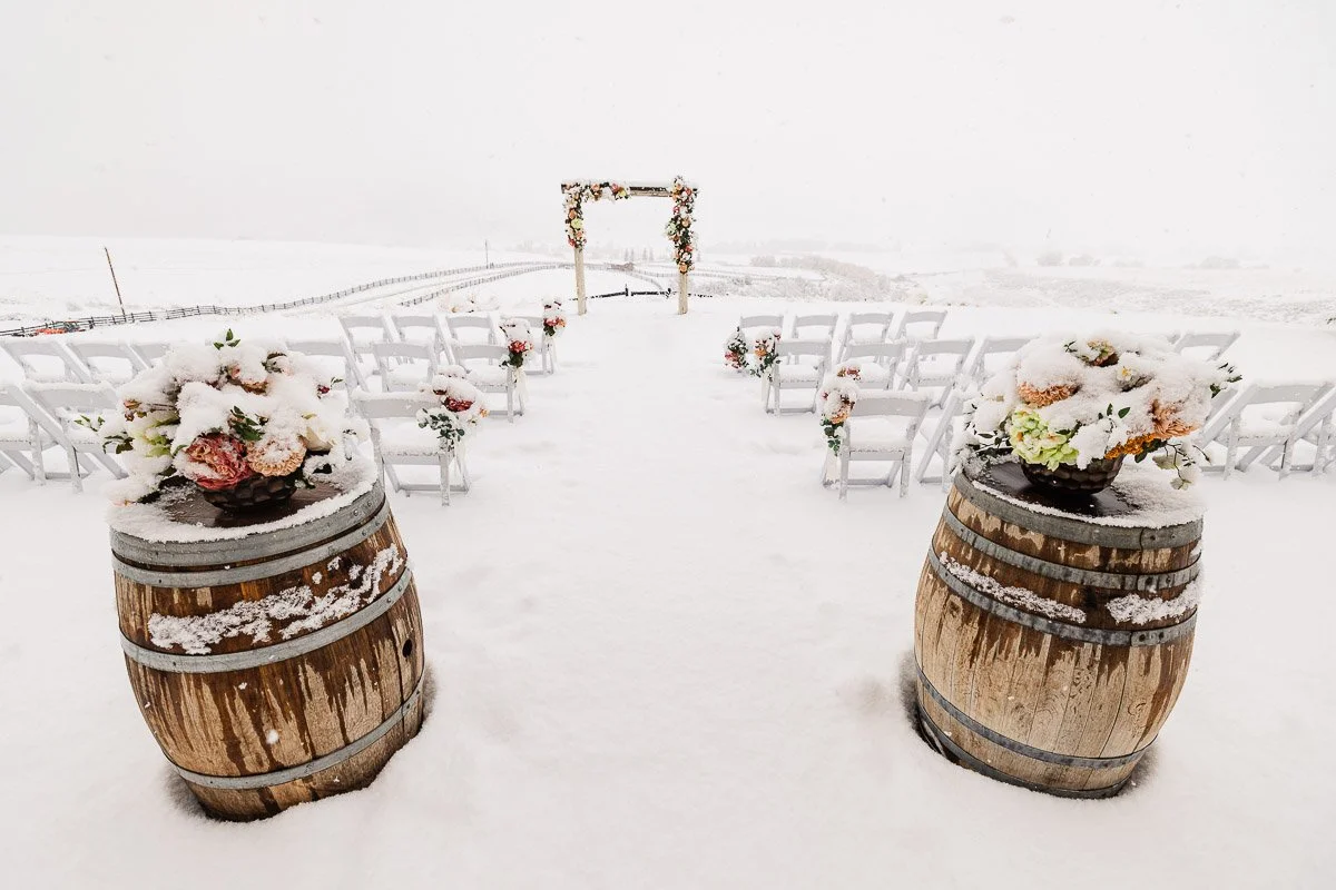 Snow-covered outdoor wedding venue with wooden barrels as aisle markers, chairs arranged in rows, and a floral arch, creating a serene winter scene.