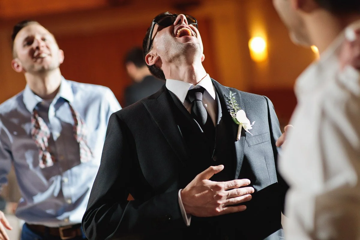 A joyful scene at a wedding reception: a man in a black suit and tie, with a boutonnière, laughs heartily while dancing, surrounded by friends.