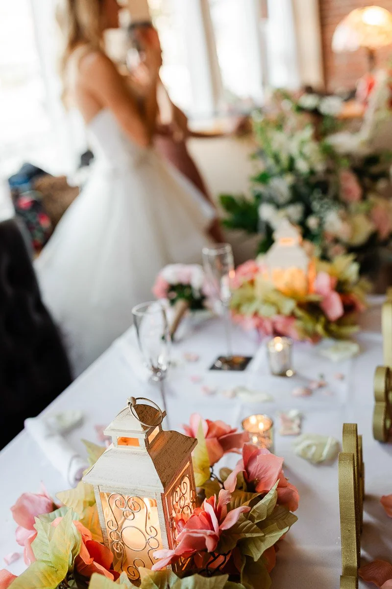 Wedding table with soft-focus bride in the background. Elegant lanterns, pink and peach flowers, and lit candles create a romantic ambiance.