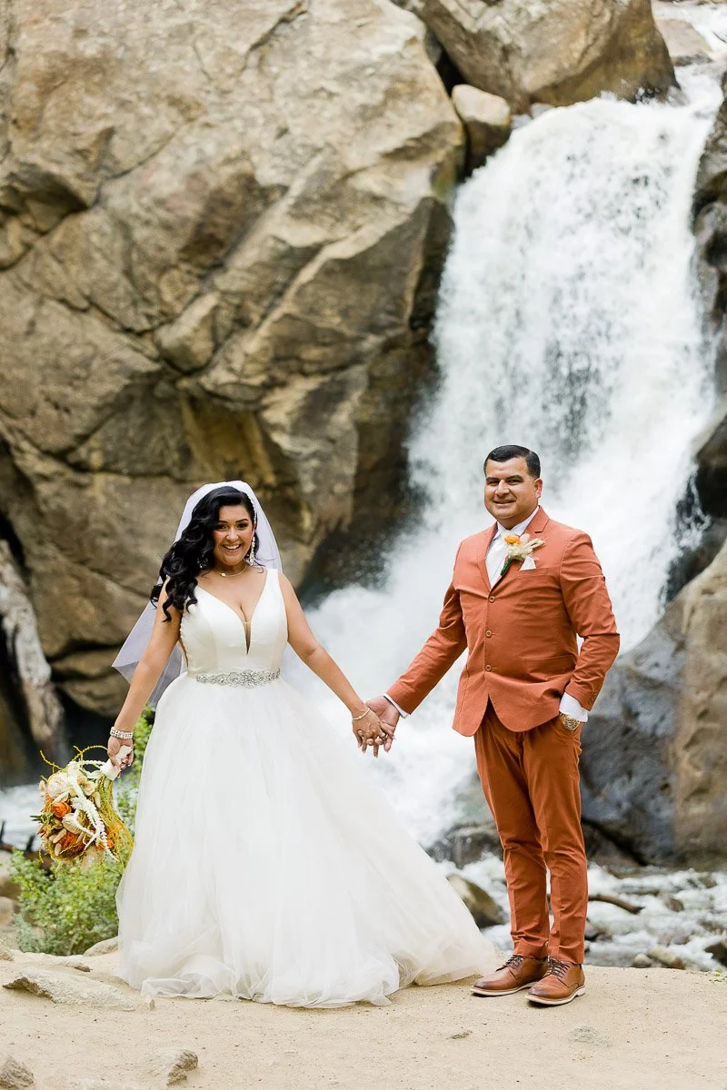Bride in a white dress and groom in an orange suit stand holding hands before a waterfall. The scene is joyful and natural, with autumn tones.