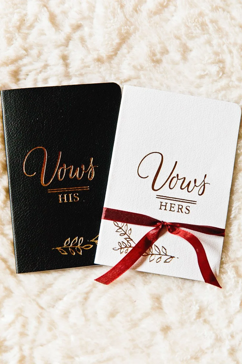 Two vow booklets lie on a soft surface; one is black with "Vows His," the other white with "Vows Hers" and a red ribbon, conveying elegance.