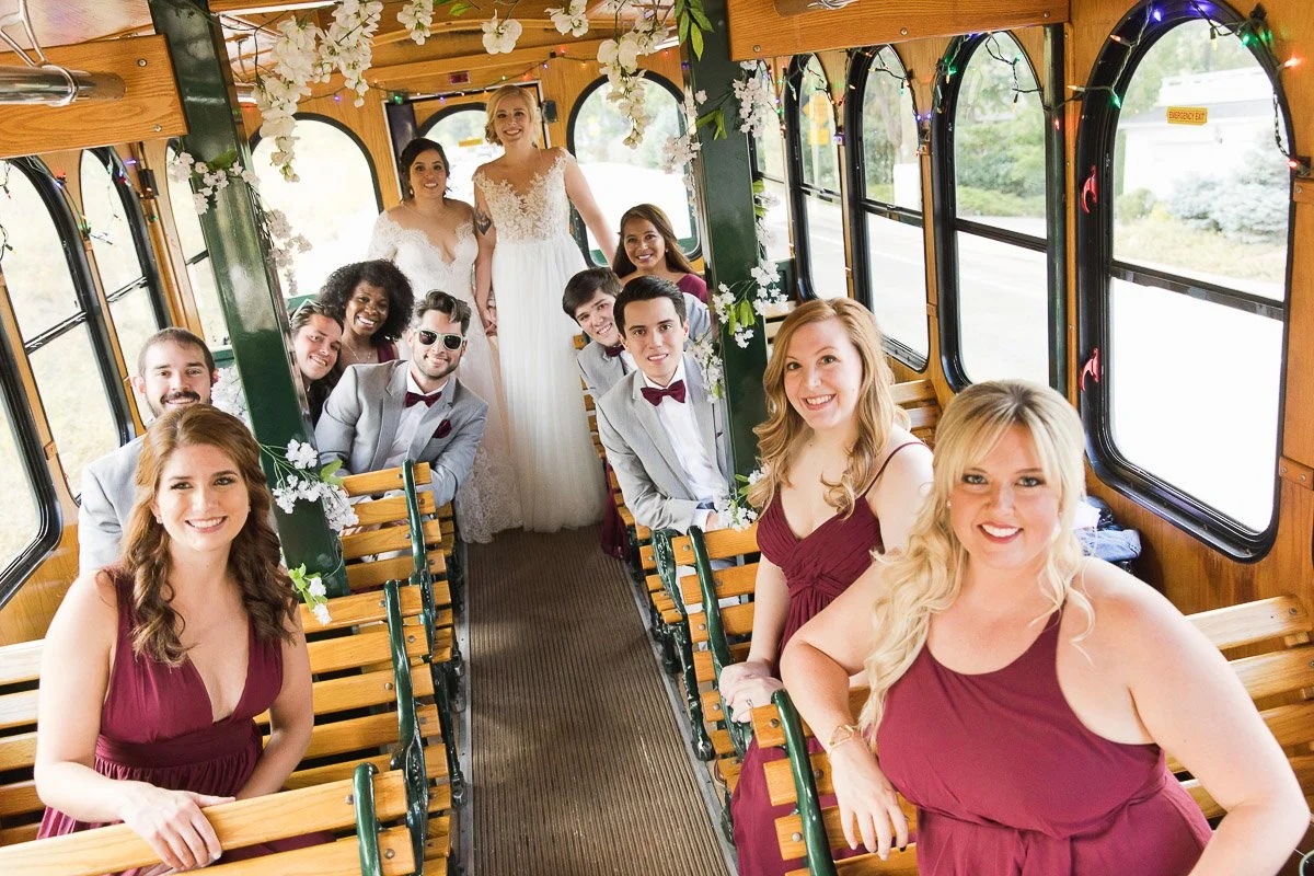 A joyful wedding party poses inside a decorated trolley. The group, in formal attire with bridesmaids in burgundy, conveys excitement and celebration.