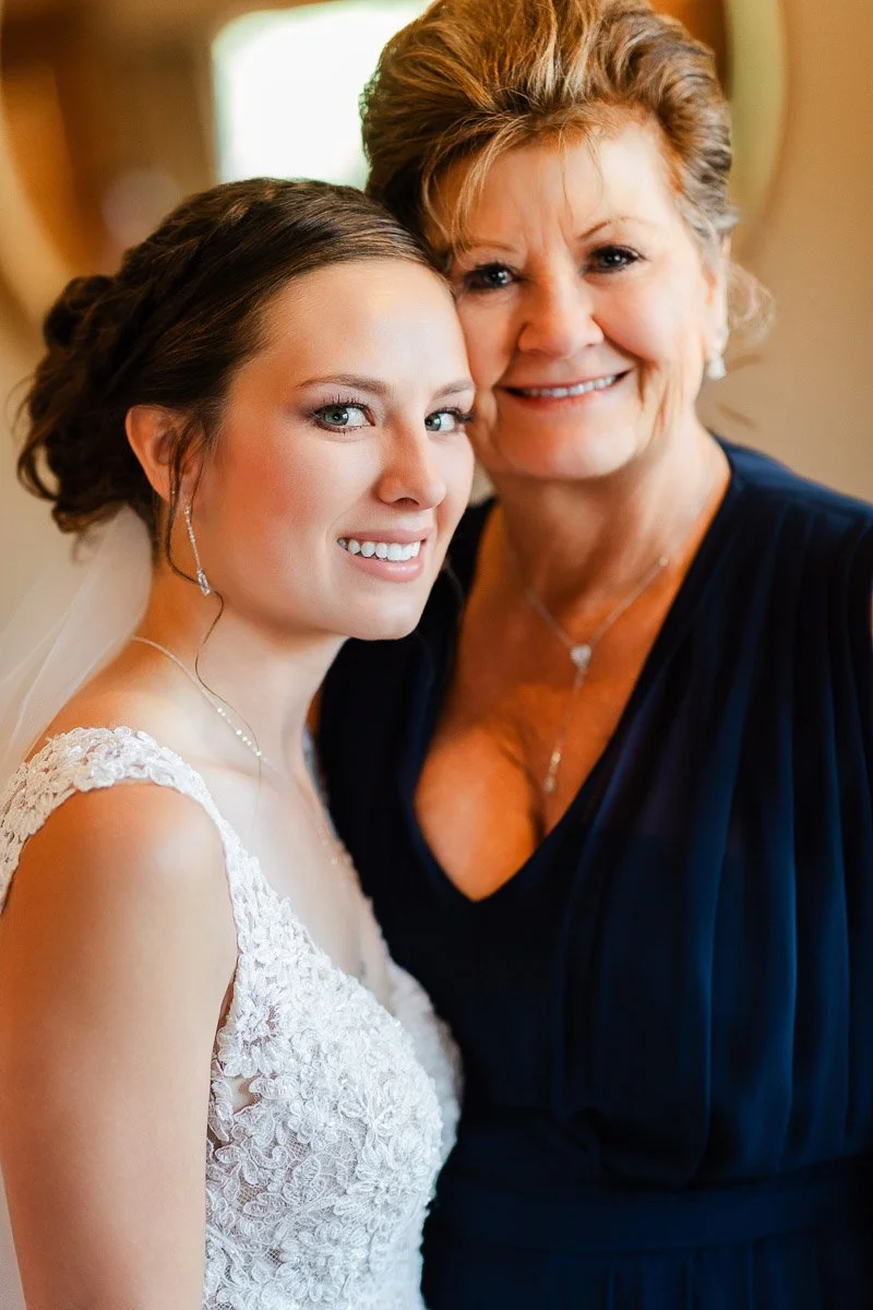 A bride in a white lace dress and veil smiles beside an older woman in a navy gown. They share a close, joyful moment in soft lighting.