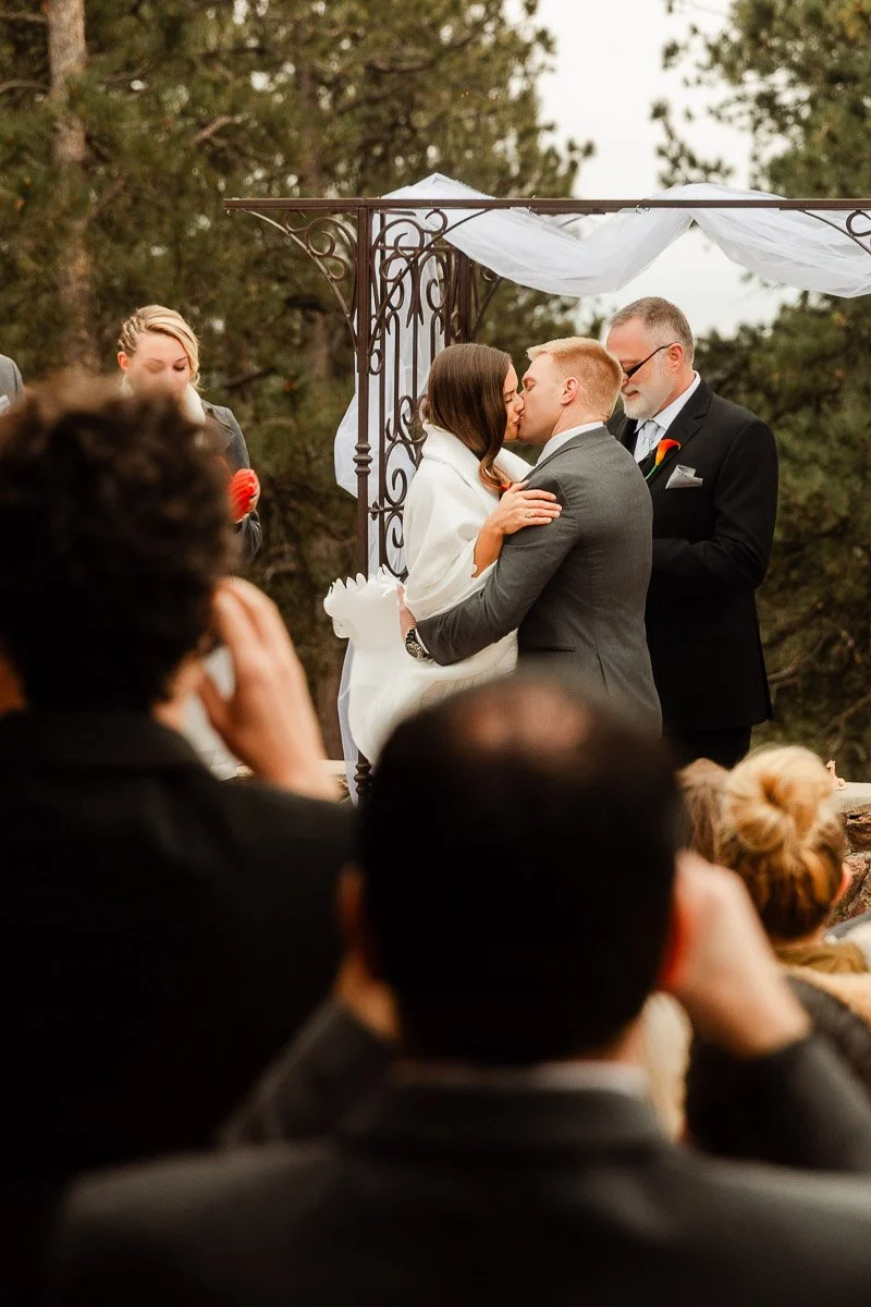 A bride and groom kiss under a metal arch with white drapery, surrounded by guests in a forest setting, creating a romantic wedding scene.
