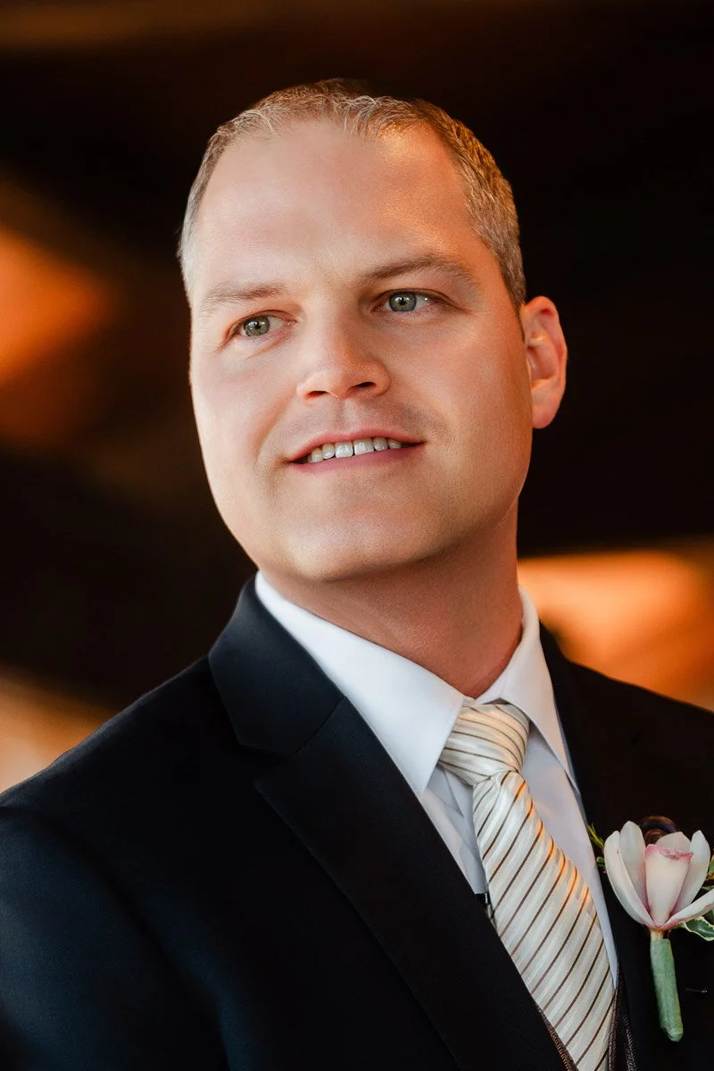Smiling man in a formal suit with a striped tie and a boutonniere. Warm, soft-focus background suggests a celebratory event.