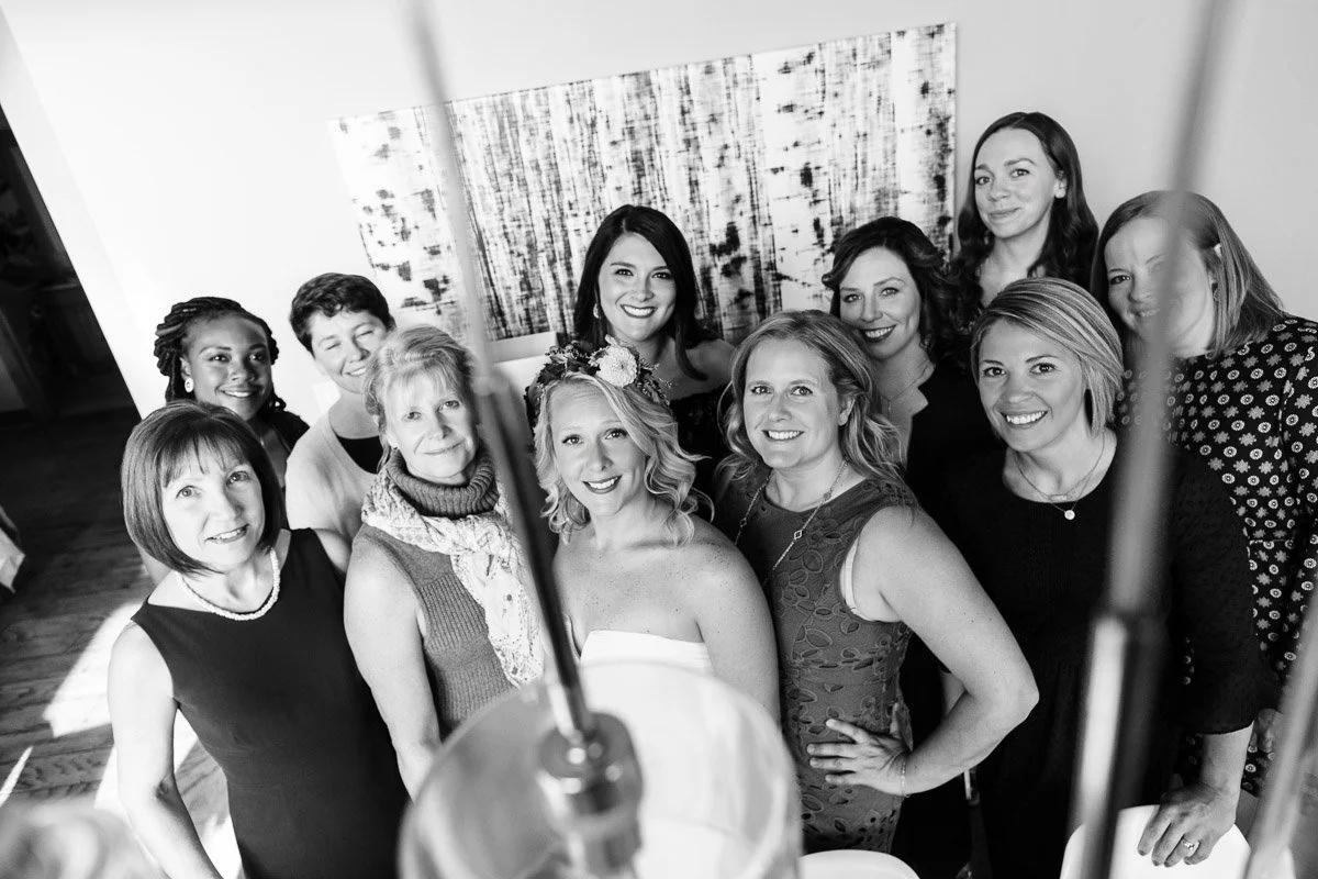 A joyful black and white photo shows a smiling bride, with a floral crown and strapless dress, surrounded by happy women in a warmly lit room.