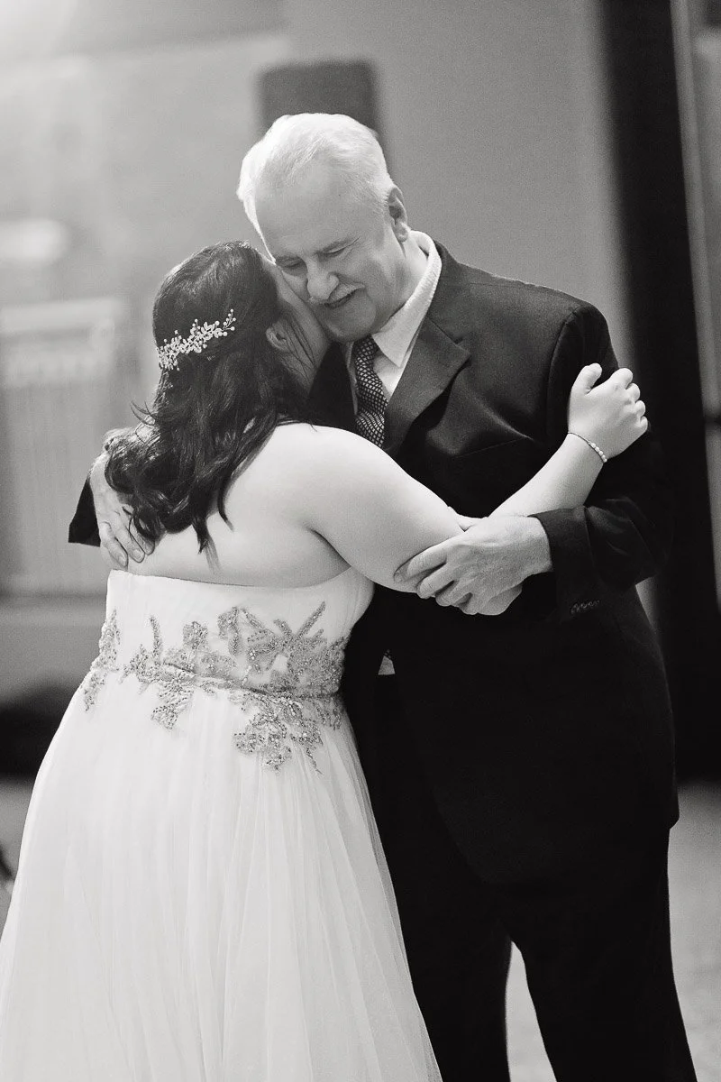 A bride in an embellished gown shares an emotional hug with an older man in a suit, likely during a wedding dance. The scene conveys warmth and affection.