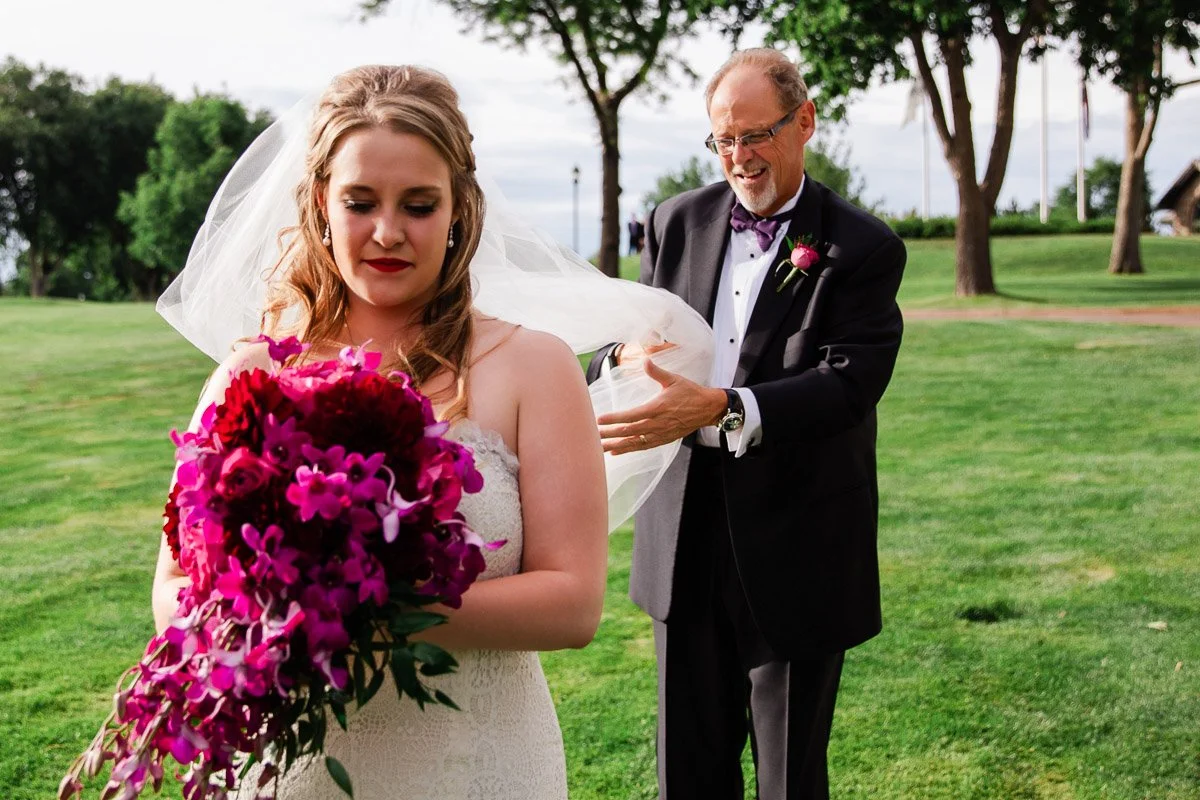 A bride with flowing hair and a vibrant bouquet stands on lush grass, eyes closed. An older man adjusts her veil, smiling warmly. Trees fill the background.