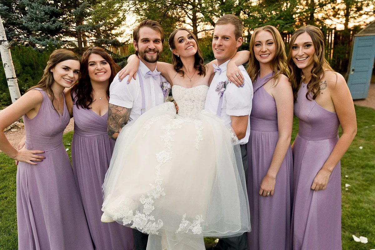 Bride in white dress, held up by two groomsmen in lavender accents, surrounded by bridesmaids in lavender dresses, all smiling joyfully outdoors.