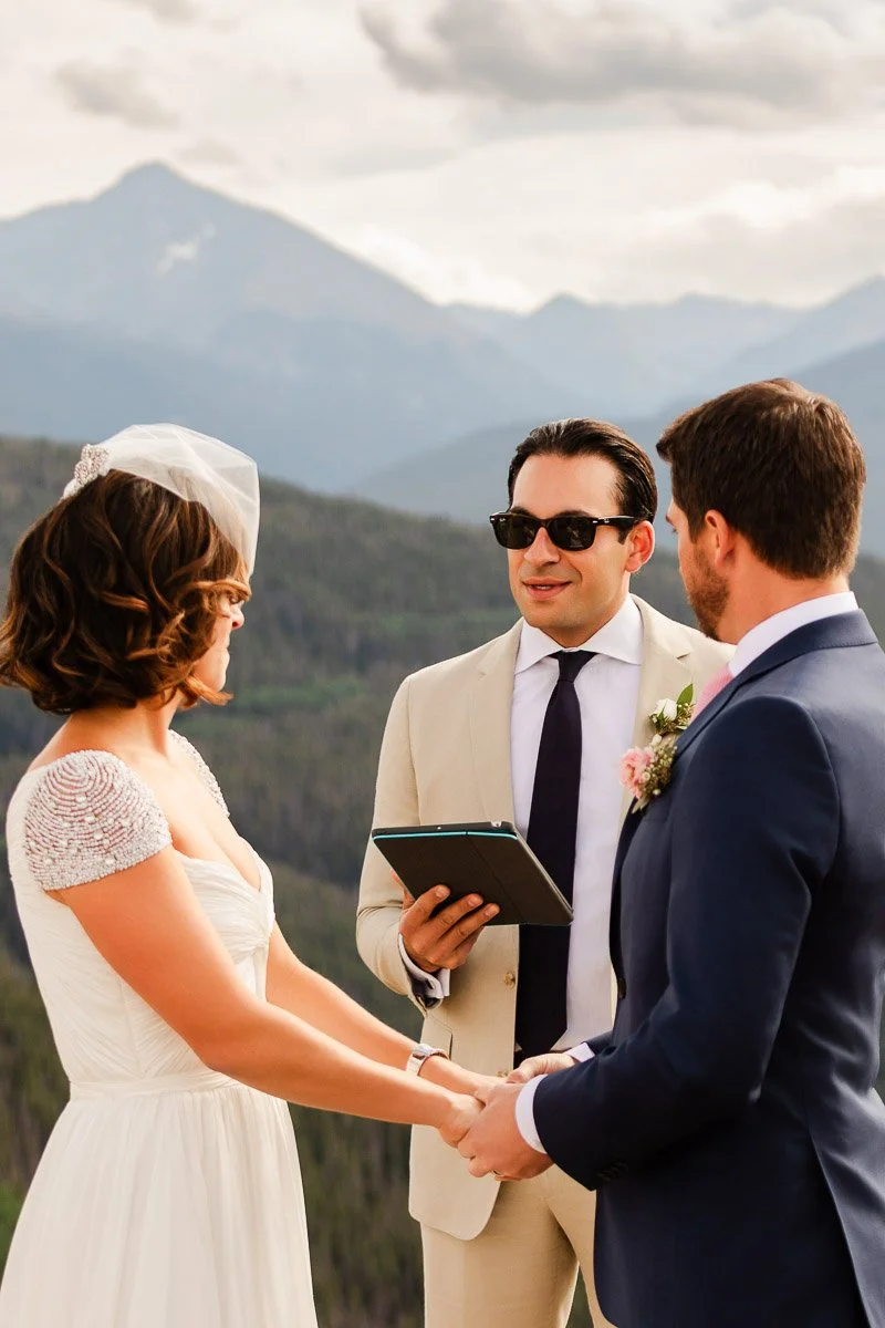 A bride and groom holding hands face each other during an outdoor mountain wedding ceremony. The officiant stands between them, smiling, under a cloudy sky.