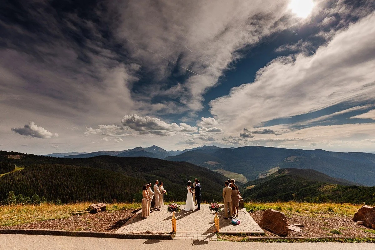 A wedding ceremony takes place on The Vail Wedding Deck, with guests in formal attire under a dramatic sky. The mood is serene and celebratory.