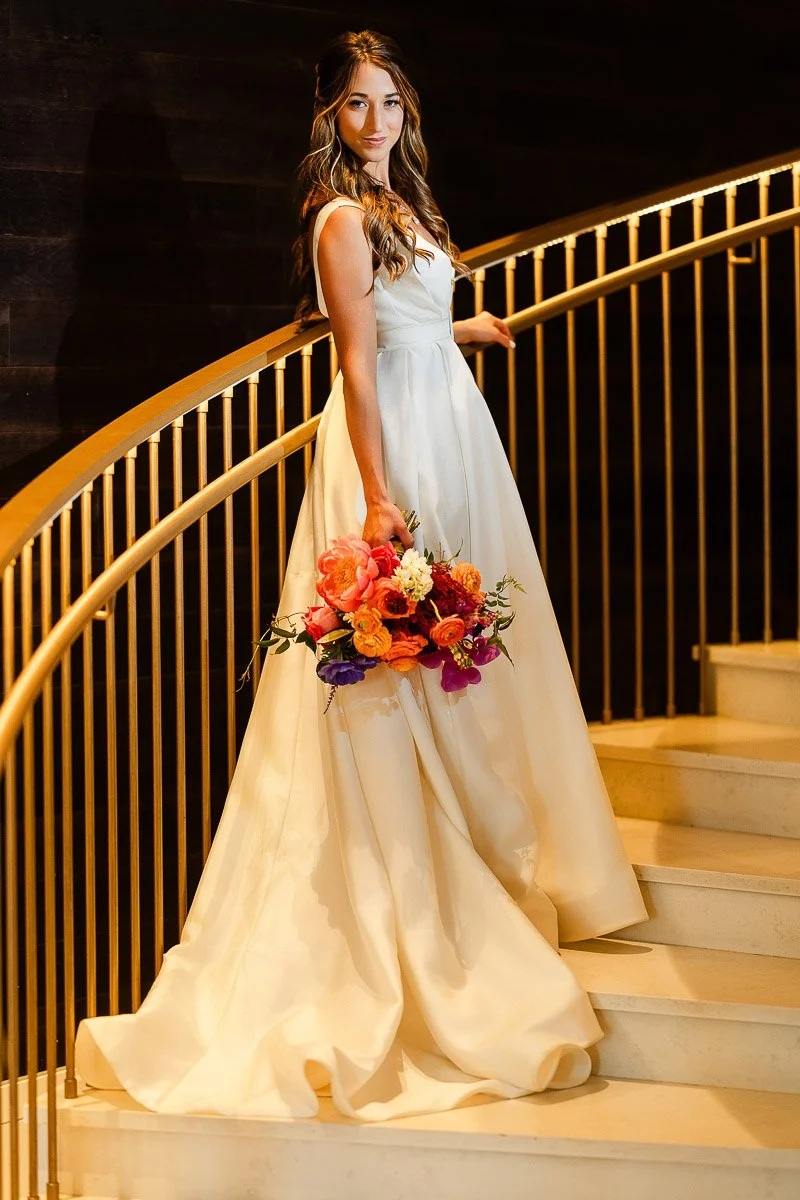Bride in elegant white dress holding vibrant bouquet stands gracefully on curved staircase with golden railing, exuding elegance and joy during an Elizabeth Hotel wedding