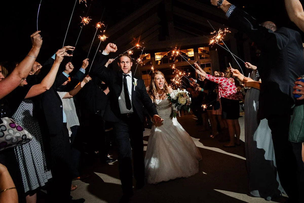 A newlywed couple joyfully runs through a crowd holding sparklers at night. The bride wears a white gown and holds flowers, capturing a festive, celebratory mood.