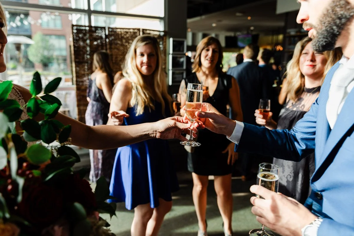 A group of well-dressed people smiling indoors during a social event. A man in a blue suit hands a glass of champagne to a woman, with sunlight streaming in.