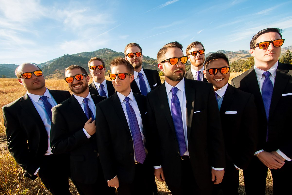 Eight men in suits and purple ties stand in a field, wearing reflective sunglasses. The mood is cheerful, with hills and a bright blue sky behind them.