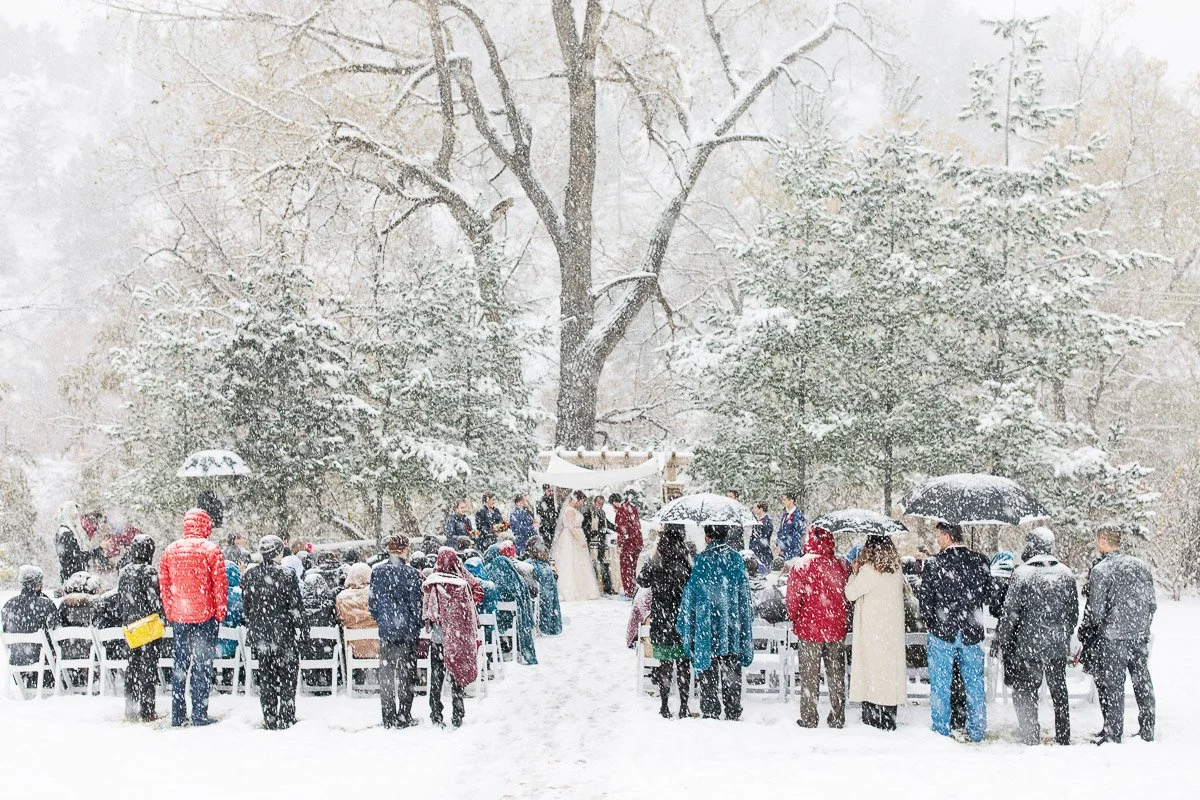 A snowy outdoor wedding ceremony with guests seated in rows under umbrellas. A couple stands under a chuppah surrounded by falling snow and tall trees.