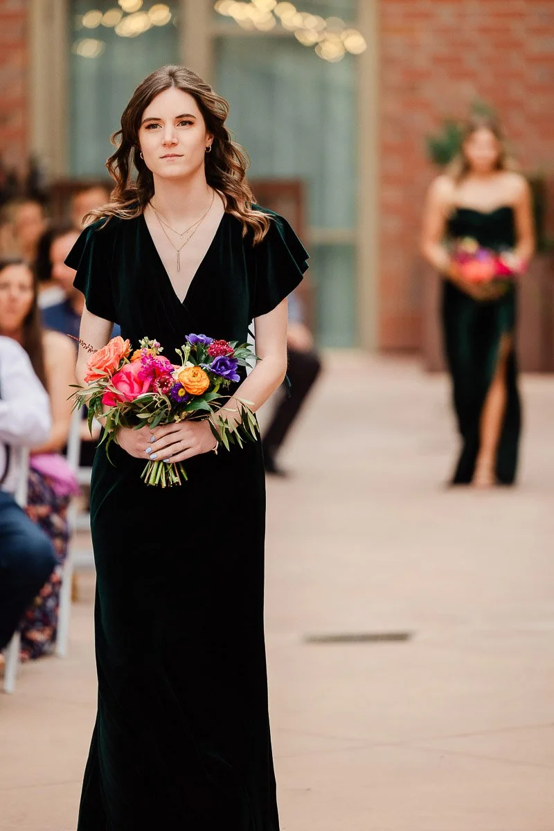 Young woman in a deep green dress walking down an aisle, holding a vibrant bouquet. Another woman follows in the background. The tone is formal and serene.