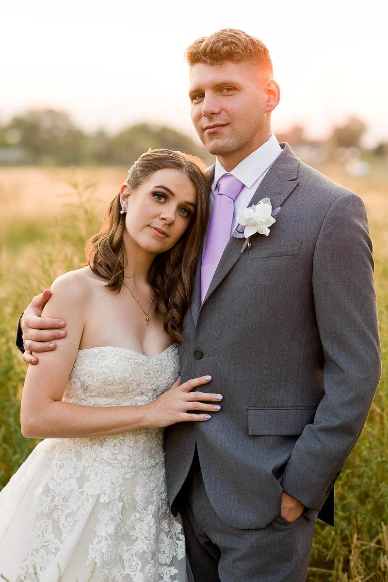 A bride and groom stand closely in a sunlit field. The bride, in a lace gown, leans on the groom, who wears a gray suit with a lavender tie, exuding love and serenity.