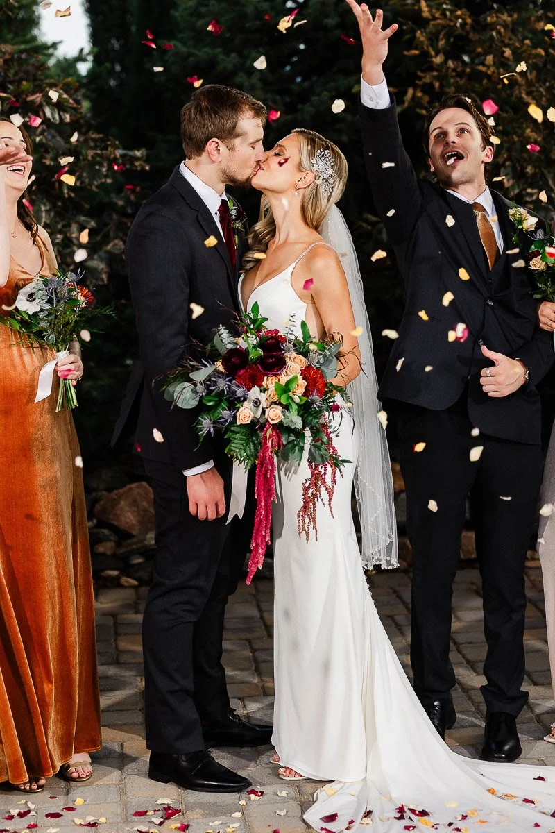 Bride and groom kiss amidst confetti and flower petals; she holds a vibrant bouquet. Joyful guests in elegant attire celebrate beside them.