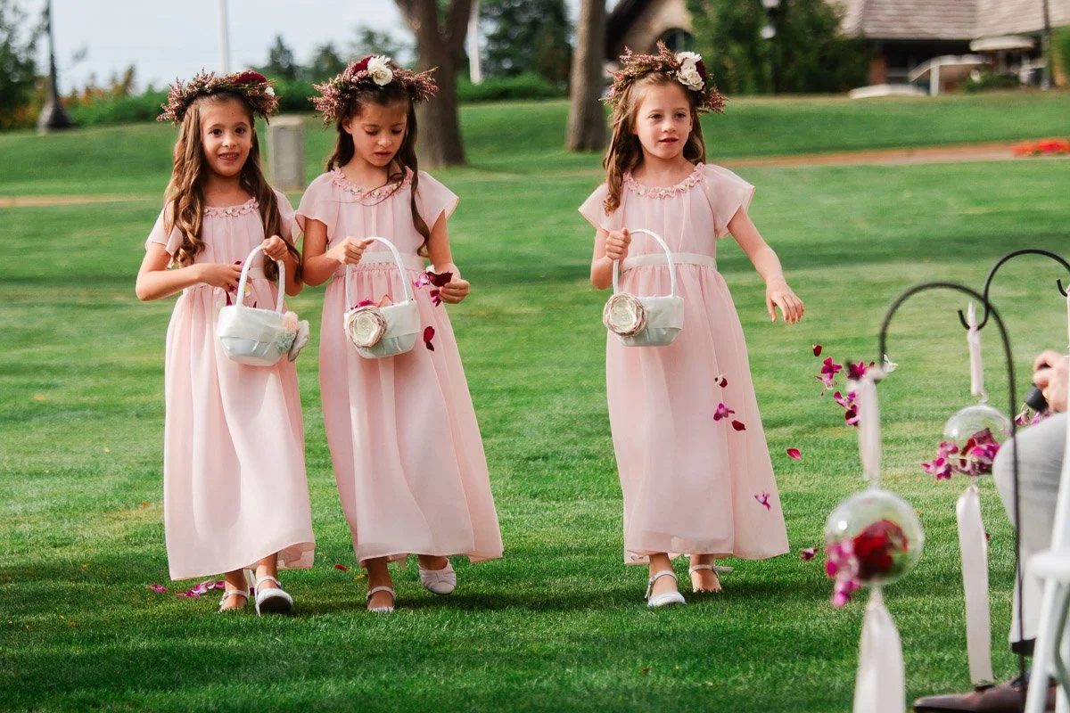 Three young flower girls in pink dresses and floral crowns walk on grass, holding white baskets and scattering petals, at an outdoor event.