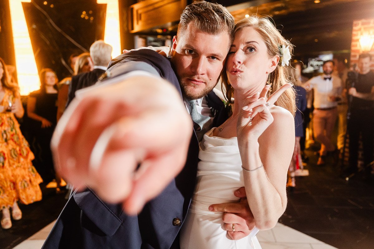 A newlywed couple at a lively wedding reception. The groom points playfully towards the camera, while the bride strikes a cheerful pose.