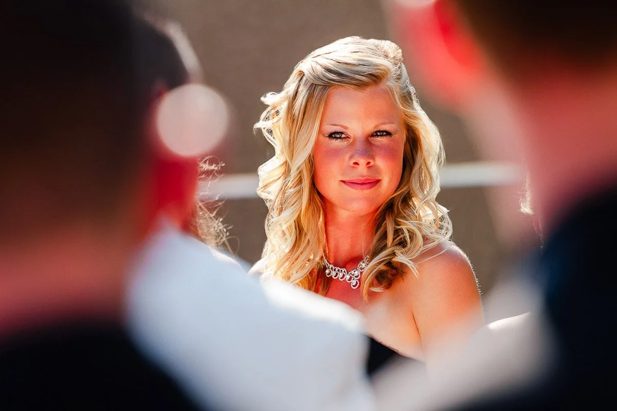 A woman with blonde, curled hair and a jeweled necklace smiles warmly in sunlight, surrounded by blurred figures, conveying a joyful, celebratory mood.