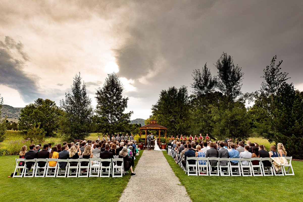 An outdoor wedding ceremony with a couple under a wooden gazebo. Dark clouds loom overhead, creating a dramatic sky. Guests sit on white chairs.