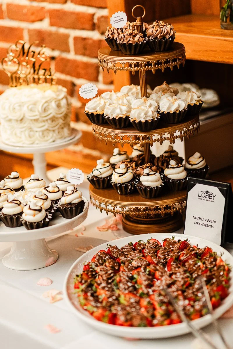 Decorative dessert table with a tiered stand of assorted cupcakes, a white frosted cake, and a platter of Nutella-drizzled strawberries.