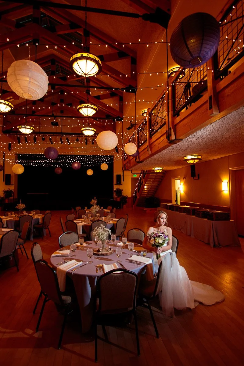 Bride in a white gown sits at a round, elegantly set table in a warmly lit reception hall decorated with string lights and paper lanterns, conveying romance.