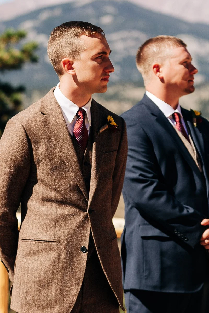 Two men in suits stand outdoors with mountains in the background. One wears a brown suit, the other navy. The scene is formal and serene.