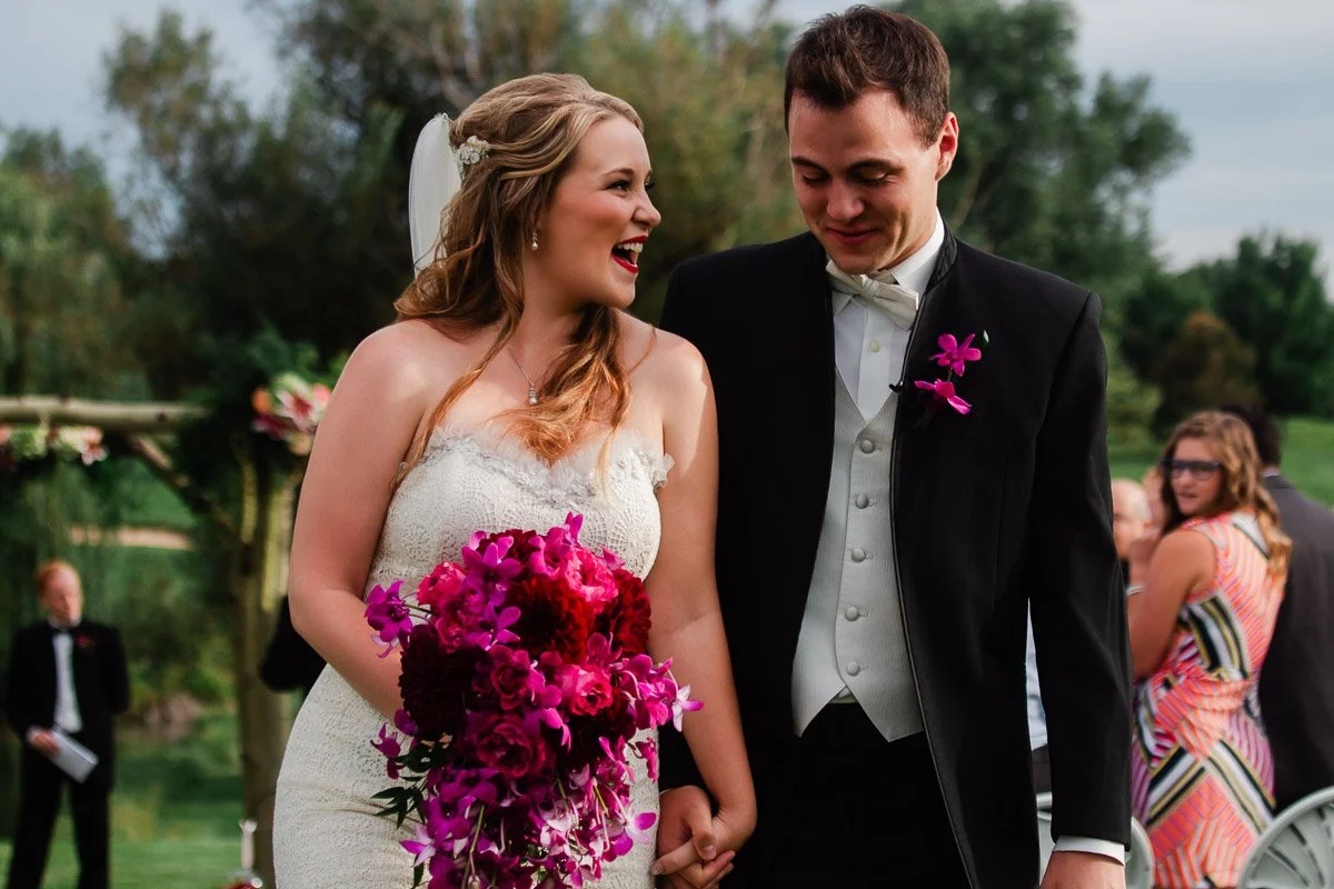 A joyful bride and groom walk hand-in-hand outdoors. She wears a lace gown and holds vibrant red and purple flowers. He is in a black tuxedo with a white vest.