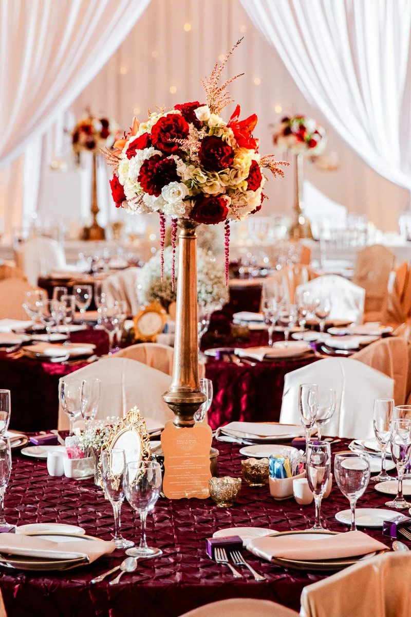 Elegant wedding reception with tall floral centerpieces featuring red and white flowers on tables draped in wine-colored cloths, creating a luxurious ambiance.