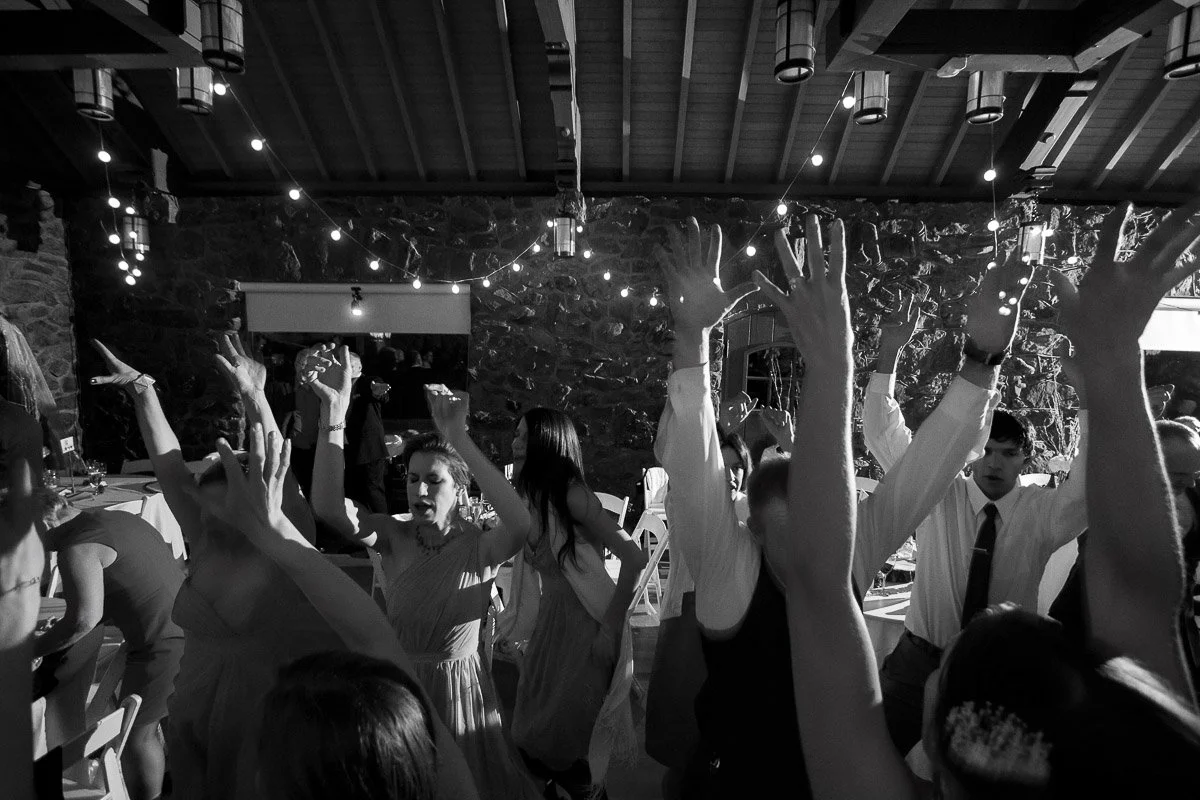 A lively black-and-white photo of people dancing with their arms raised under string lights at an indoor party, conveying joy and celebration.