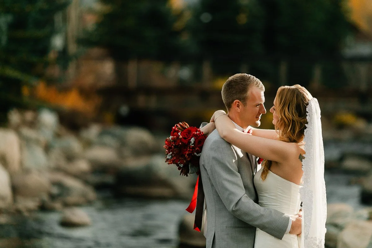A bride and groom embrace by a serene river. The bride holds red flowers and wears a veil, while the groom is in a gray suit. The scene is warm and romantic.