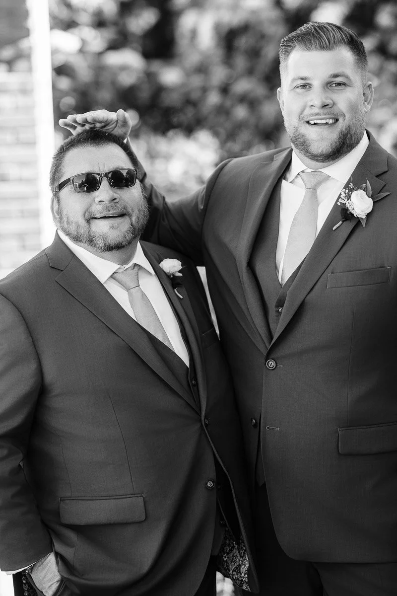 Two men in suits, smiling warmly. One pats the other's head playfully. They wear boutonnières, suggesting a wedding setting. Black and white photo.